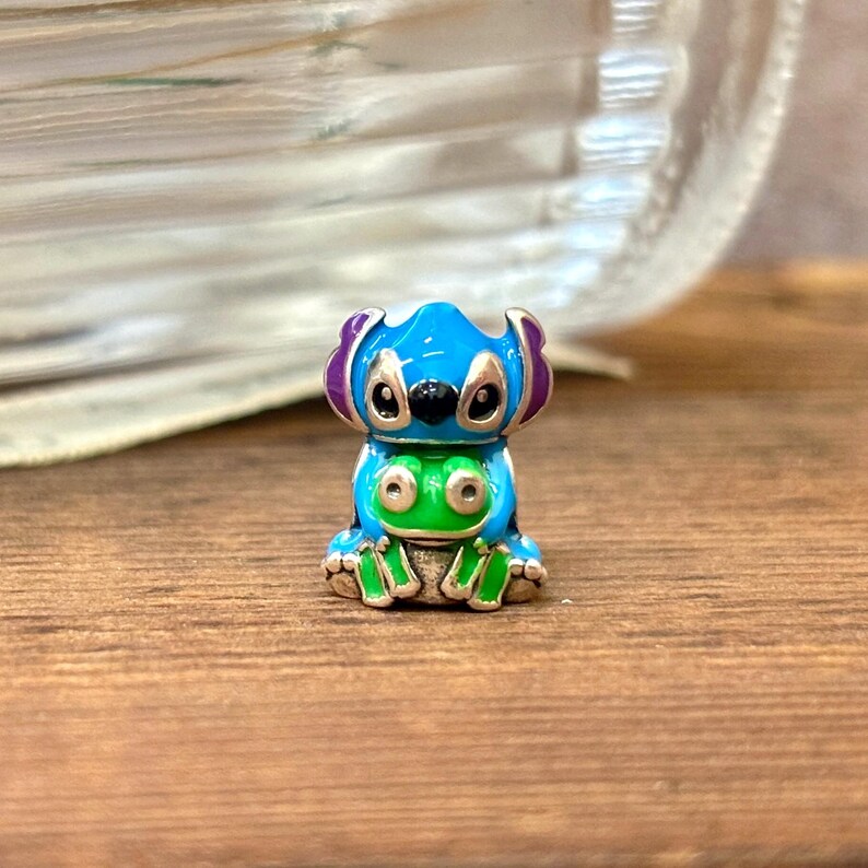 May include: A small, colourful figurine of a cartoon character. The figure is primarily blue with purple ears, black eyes, and a green belly. The figurine is sitting on a wooden surface.