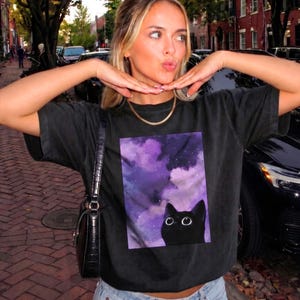 Puede incluir: Una camiseta negra de manga corta con un estampado gráfico rectangular de un gato negro con ojos grandes sobre un cielo nocturno nublado de color morado y blanco. La camiseta se combina con un bolso de hombro negro y un collar de oro.