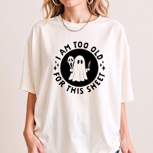 Puede incluir: Una camiseta de manga corta color blanco roto con un gráfico circular negro en el pecho. El gráfico muestra un fantasma blanco sosteniendo una máscara y un cuchillo, rodeado por el texto negro "I AM TOO OLD FOR THIS SHEET" y estrellas.