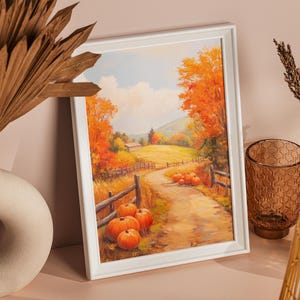 Puede incluir: Un marco blanco rectangular que contiene una pintura de paisaje otoñal con un camino de tierra, calabazas naranjas, cercas de madera y árboles de colores vibrantes bajo un cielo nublado. La obra se exhibe junto a follaje seco decorativo.