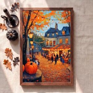 Puede incluir: Una impresión de arte enmarcada que representa una escena callejera otoñal con árboles naranjas, luces de cadena brillantes y un mercado concurrido. La pintura muestra una gran calabaza naranja en primer plano, edificios de piedra y hojas caídas.