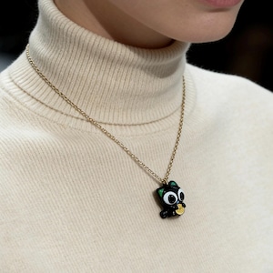 May include: A gold chain necklace with a black cat pendant. The cat pendant has large white eyes, green ears, and is holding a gold coin. The person is wearing a cream-colored turtleneck sweater.