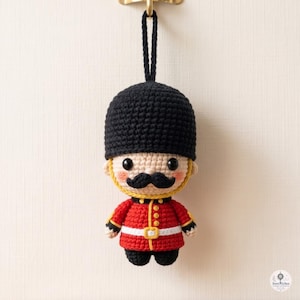 May include: A crocheted amigurumi figure of a British royal guard wearing a tall black bearskin hat, a red tunic with gold trim, a white belt, and black trousers. The figure has a beige face, black eyes, and a prominent black mustache.