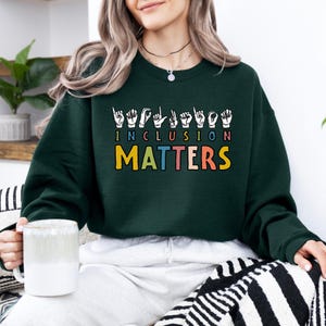 May include: A dark green sweatshirt with the words "INCLUSION MATTERS" in a colorful design. The word "INCLUSION" is above "MATTERS" and is written in sign language symbols. The sweatshirt is a crew neck style.