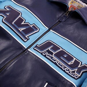K&ouml;nnte beinhalten: Marineblaue Lederjacke mit hellblauen und wei&szlig;en Akzenten. Die Jacke tr&auml;gt den Schriftzug "AV8" und "REX NITRO RUN" in einem stilisierten Schriftzug. Sie hat einen Rei&szlig;verschluss und einen Kragen.