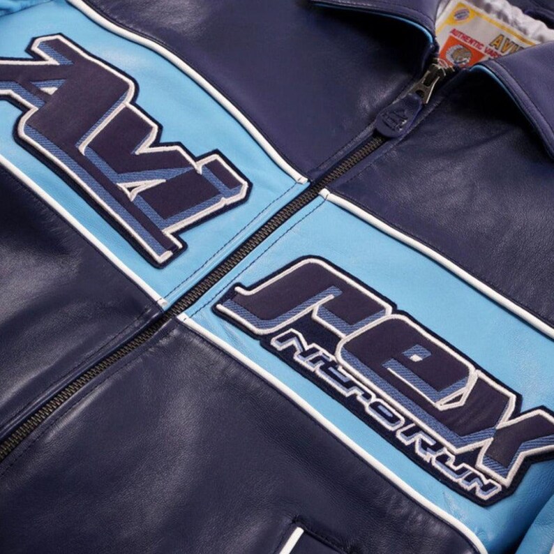 K&ouml;nnte beinhalten: Marineblaue Lederjacke mit hellblauen und wei&szlig;en Akzenten. Die Jacke tr&auml;gt den Schriftzug "AV8" und "REX NITRO RUN" in einem stilisierten Schriftzug. Sie hat einen Rei&szlig;verschluss und einen Kragen.