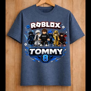 May include: Blue t-shirt featuring a Roblox graphic with multiple characters. The shirt has the text "ROBLOX" in white and red, and "TOMMY 8" in blue and white. The shirt is hanging on a wooden hanger.