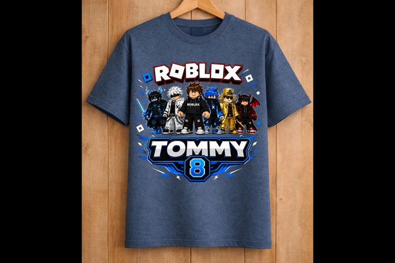 May include: Blue t-shirt featuring a Roblox graphic with multiple characters. The shirt has the text "ROBLOX" in white and red, and "TOMMY 8" in blue and white. The shirt is hanging on a wooden hanger.