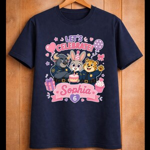 May include: Navy blue t-shirt featuring cartoon characters from Zootopia. The design includes the text "Let's Celebrate!" and "Sophia 6" with a cake, balloons, and gifts. The shirt is hanging on a wooden hanger.