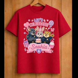 May include: Red t-shirt featuring a colorful graphic with the text "Let's Celebrate!" and the name "Sophia" with the number "6". The design includes cartoon characters, balloons, a cake, and a cupcake.