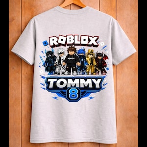 May include: A light gray t-shirt featuring a Roblox design with several cartoon characters. The shirt has the word "ROBLOX" in red and white, and the name "TOMMY" with the number "8" in blue. The shirt is hanging on a wooden hanger.