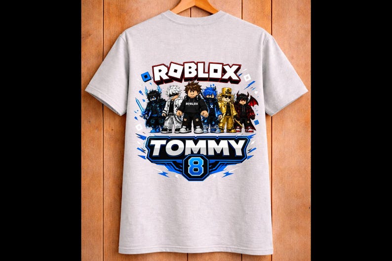 May include: A light gray t-shirt featuring a Roblox design with several cartoon characters. The shirt has the word "ROBLOX" in red and white, and the name "TOMMY" with the number "8" in blue. The shirt is hanging on a wooden hanger.
