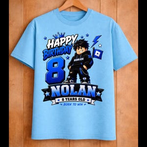 May include: Light blue t-shirt with a graphic design for an 8-year-old's birthday. The design includes the text "Happy Birthday," the number "8," the name "Nolan," and a Roblox character. The shirt also says "9 years old" and "Born to Win."