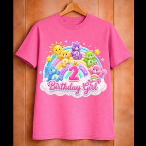 Peut inclure: T-shirt rose avec un motif coloré de personnages d'ours de dessin animé, un arc-en-ciel et le texte "Birthday Girl" avec le chiffre "2". Le motif comprend un soleil, des étoiles et des nuages. Le t-shirt est suspendu à un cintre en bois.