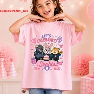 May include: A light pink t-shirt featuring a colorful graphic of cartoon characters, a cake, and the text "LET'S CELEBRATE!" and "Sophia 6". The shirt is short-sleeved and suitable for a birthday celebration.