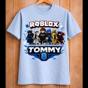 May include: Light blue Roblox-themed t-shirt featuring a group of cartoon characters. The shirt has the word "ROBLOX" in red and white, with the name "TOMMY" and the number "8" in blue and white.