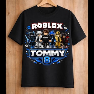 May include: Black t-shirt featuring the word "ROBLOX" in white and blue, with cartoon characters. Below, the name "TOMMY" and the number "8" are displayed in a blue and white design. The shirt is hanging on a wooden hanger.