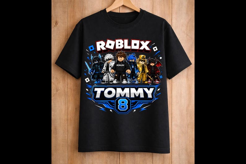 May include: Black t-shirt featuring the word "ROBLOX" in white and blue, with cartoon characters. Below, the name "TOMMY" and the number "8" are displayed in a blue and white design. The shirt is hanging on a wooden hanger.