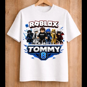 May include: White t-shirt featuring a colorful graphic of Roblox characters. The design includes the word "ROBLOX" in red and white, the name "TOMMY" in blue, and the number "8". The shirt is hanging on a wooden hanger.