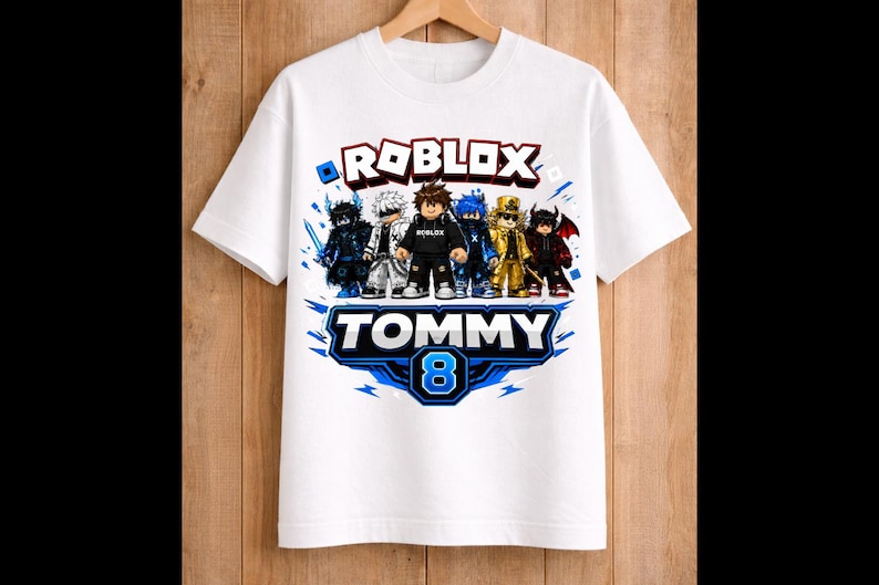 May include: White t-shirt featuring a colorful graphic of Roblox characters. The design includes the word "ROBLOX" in red and white, the name "TOMMY" in blue, and the number "8". The shirt is hanging on a wooden hanger.