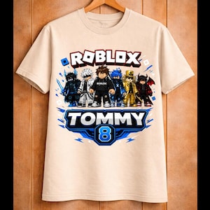 May include: Beige t-shirt featuring a Roblox design with several cartoon characters. The shirt has the text "ROBLOX" above a banner that reads "TOMMY 8" in blue and white. The shirt is hanging on a wooden hanger.
