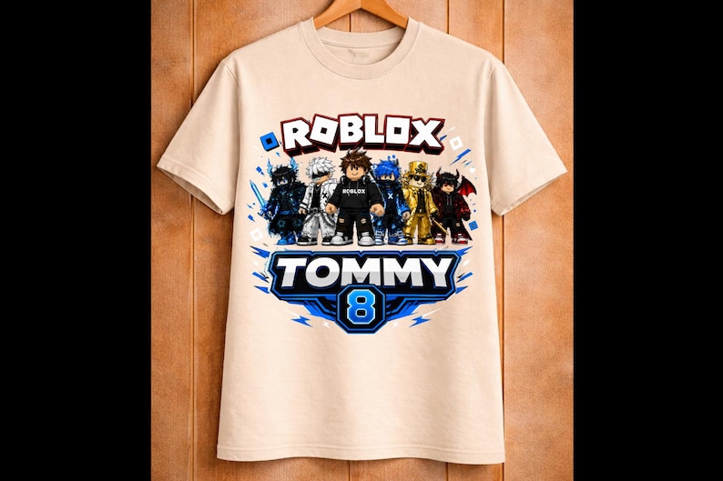 May include: Beige t-shirt featuring a Roblox design with several cartoon characters. The shirt has the text "ROBLOX" above a banner that reads "TOMMY 8" in blue and white. The shirt is hanging on a wooden hanger.
