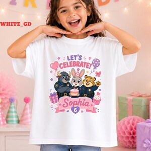 May include: White t-shirt featuring a colorful graphic of cartoon characters, a cake, and the text "LET'S CELEBRATE!" and "Sophia 6". The design includes a heart, stars, and a balloon. The shirt is worn by a young girl.