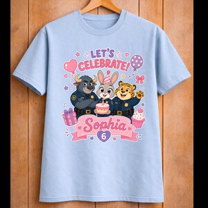 May include: Light blue t-shirt featuring a colorful graphic with three cartoon animal characters in police uniforms. The design includes the text "LET'S CELEBRATE!" and "Sophia 6" with birthday-themed elements like balloons, a cake, and cupcakes.