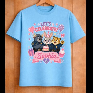 May include: Light blue t-shirt featuring a birthday design with cartoon characters. The shirt has the text "LET'S CELEBRATE!" and the name "Sophia" with the number "6". Includes images of a cake, balloons, and cupcakes.