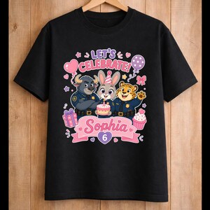 May include: Black t-shirt featuring a colorful graphic with the text "Let's Celebrate!" and the name "Sophia" with the number "6". The design includes cartoon characters, balloons, and a cupcake.