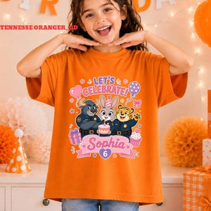 May include: Orange t-shirt featuring a colorful graphic with the text "LET'S CELEBRATE!" and the name "Sophia" with the number "6". The graphic includes cartoon characters, balloons, and a cupcake.