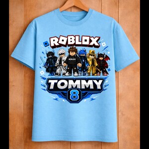 May include: Light blue t-shirt featuring a Roblox graphic with several cartoon characters. The shirt has the text "ROBLOX" in red and white, and "TOMMY 8" in blue and white. The shirt is hanging on a wooden hanger.