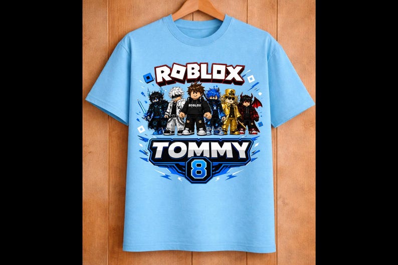 May include: Light blue t-shirt featuring a Roblox graphic with several cartoon characters. The shirt has the text "ROBLOX" in red and white, and "TOMMY 8" in blue and white. The shirt is hanging on a wooden hanger.
