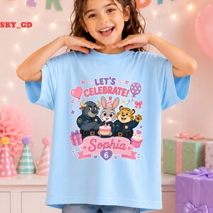 May include: A light blue t-shirt featuring a colorful graphic of cartoon characters from Zootopia, with the text "LET'S CELEBRATE" and the name "Sophia" with the number "6". The design includes a cake, balloons, and gift boxes.