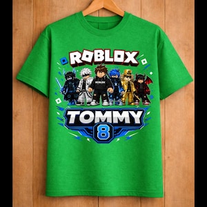 May include: Green t-shirt featuring the word "ROBLOX" in white and red, with a group of cartoon characters. Below, the name "TOMMY" and the number "8" are displayed in a blue and white design. The shirt is hanging on a wooden hanger.