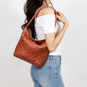 May include: A woven leather shoulder bag in a warm brown hue. The bag features a braided strap and a spacious, slouchy design. The texture of the leather is visible, showcasing the intricate weave pattern. The bag is a stylish accessory.