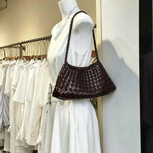 Puede incluir: Un bolso de hombro de cuero trenzado marrón oscuro con una correa trenzada. El bolso se exhibe en un maniquí blanco, mostrando su forma rectangular y su intrincado patrón de tejido. El color del bolso contrasta con la ropa y el fondo blancos.