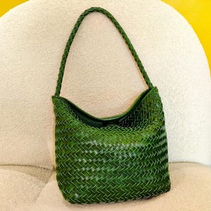 Peut inclure: Un sac à bandoulière vert en cuir tressé avec une poignée tressée. Le sac a une forme incurvée et une surface texturée, posé sur une chaise crème et un fond jaune.