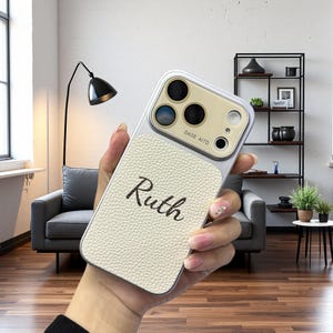 Può includere: Una custodia per telefono personalizzata con il nome "Ruth" in corsivo. La custodia ha un pannello color crema, strutturato, simile alla pelle e una cornice argentata. L'area della fotocamera ha una finitura dorata. La custodia è tenuta in mano.