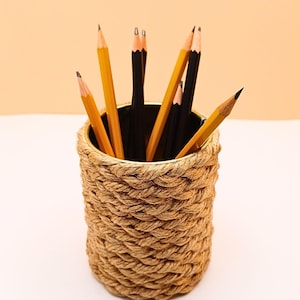 Può includere: Un portapenne cilindrico avvolto in corda color naturale, pieno di matite gialle e nere. Le matite sono temperate e pronte all'uso. Il portapenne è appoggiato su una superficie bianca contro uno sfondo color pesca.