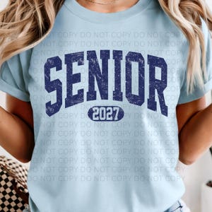 K&ouml;nnte beinhalten: Hellblaues T-Shirt mit dem Wort "SENIOR" in marineblauen, verwitterten Blockbuchstaben und der Jahreszahl "2027" darunter. Das Shirt hat einen Rundhalsausschnitt und kurze &Auml;rmel.