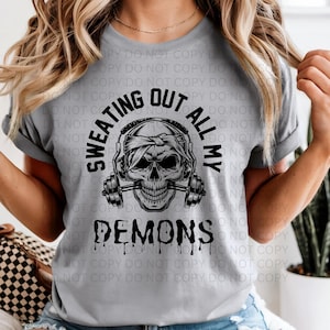 Może przedstawiać: Jasnoszara koszulka z nadrukiem czaszki w słuchawkach i trzymającej sztangi. Tekst "SWEATING OUT ALL MY DEMONS" jest wydrukowany w czarnym, ociekającym kroplami kroju pisma, tworząc odważny projekt graficzny.