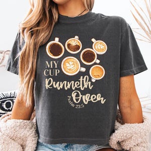 May include: A charcoal gray short-sleeve t-shirt featuring a graphic of six coffee cups in various styles. The text reads "MY CUP RUNNETH OVER" and "Psalm 23:5" in a cream-colored font. The design is centered on the front of the garment.