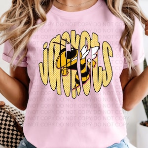 May include: A light pink t-shirt featuring the word "JACKETS" in a yellow, bubble-style font with a black outline. A cartoon bee with yellow and black stripes is integrated into the design, adding a playful touch to the apparel.