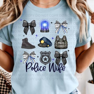 以下が含まれることがあります： 警察をテーマにしたグラフィックが特徴のライトブルーのTシャツ。デザインには、警察のバッジ、警察の帽子、警察のベスト、リボン、そして「Police Wife」の文字が含まれています。