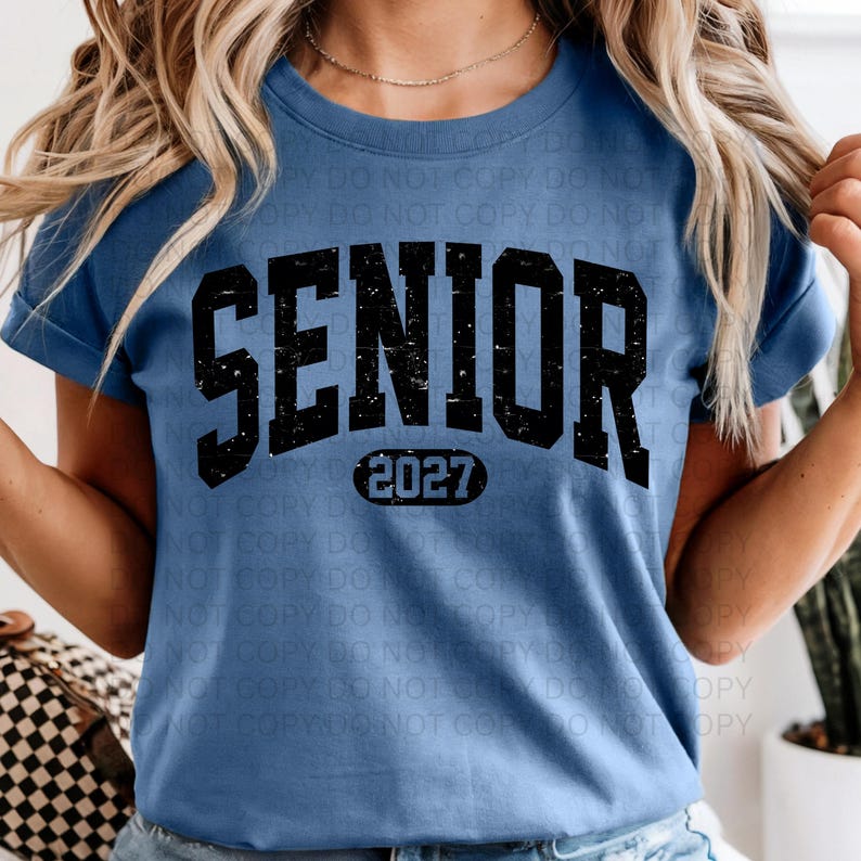 K&ouml;nnte beinhalten: Hellblaues T-Shirt mit dem Wort "SENIOR" in gro&szlig;en, verwaschenen schwarzen Buchstaben. Darunter befindet sich die Zahl "2027" in einem schwarzen Oval. Das T-Shirt hat kurze &Auml;rmel.