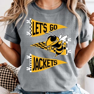 Peut inclure: T-shirt gris avec un graphisme jaune, noir et blanc d'une mascotte d'abeille et des fanions qui disent "LET'S GO" et "JACKETS". L'abeille sourit et tient un drapeau à damier. Le t-shirt a des manches courtes.