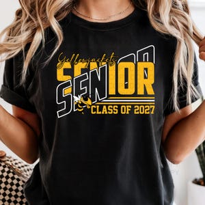 Könnte beinhalten: Schwarzes T-Shirt mit gelb-weißem Aufdruck. Der Aufdruck lautet "Yellowjackets SENIOR CLASS OF 2027" mit einer Bienenillustration. Das T-Shirt hat hochgekrempelte Ärmel.