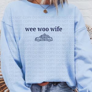 Può includere: Felpa azzurra con la scritta "wee woo wife" e una grafica di un'auto della polizia blu navy. La felpa è a girocollo e sembra essere realizzata in un materiale morbido.
