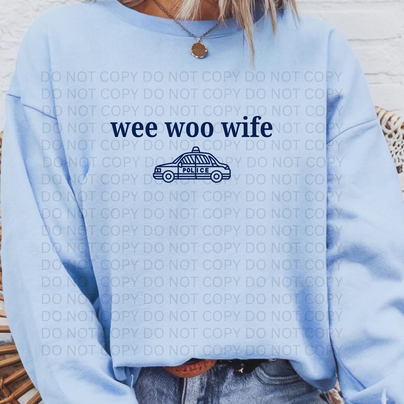 May include: Light blue sweatshirt with the text "wee woo wife" and a police car graphic in navy blue. The sweatshirt is a crew neck style and appears to be made of a soft material.